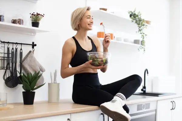 Une femme souriante en tenue de sport mange une salade fraîche. Elle savoure son repas sain dans une cuisine lumineuse.