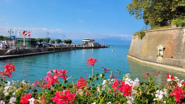 Fiori rossi e bianchi incorniciano il sirmione lungolago, con il molo, il battello e le montagne sullo sfondo.