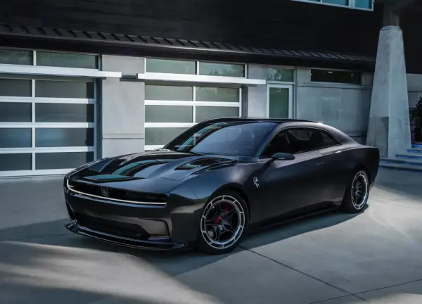 Nowoczesny, ciemnoszary Dodge Daytona z charakterystycznym pasem LED z przodu, zaparkowany przed nowoczesnym budynkiem z garażem.