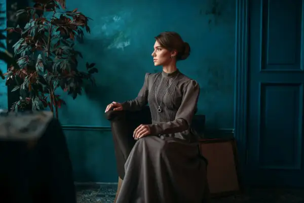 Elegancka kobieta w sukni w stylu dark academia siedzi w fotelu, wpatrzona w dal. W tle ciemnozielona ściana i rośliny.