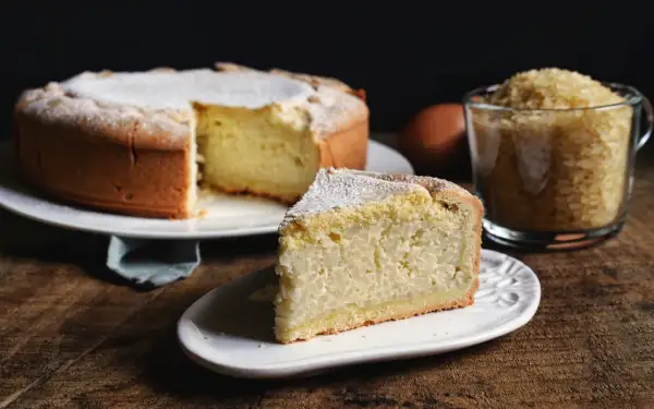 Una fetta di torta di riso, la ricetta originale, cosparsa di zucchero a velo, accanto alla torta intera e a una tazza di riso.