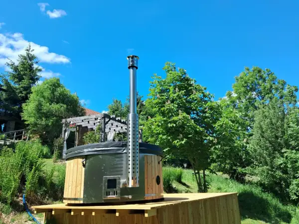 Drewniane jacuzzi w ziemi z metalowym kominem, otoczone zielenią i błękitnym niebem. Idealne miejsce na relaks.