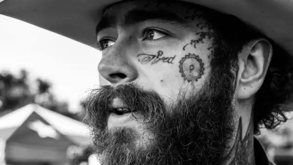 Zbliżenie na twarz Post Malone'a w czerni i bieli, z tatuażem "Tired" na policzku. Fani w Polsce czekają na jego koncert.