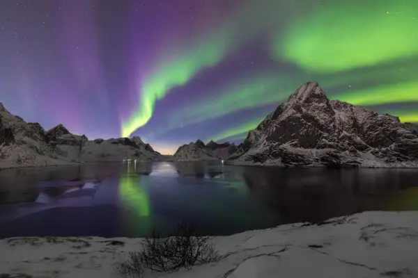 Spectaculaire foto's van het noorderlicht boven besneeuwde bergen en een bevroren meer. De groene en paarse gloed reflecteert in het water.