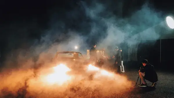 A film crew uses artificial light examples, including a bright spotlight and car headlights, to create a smoky, dramatic scene with men in hats gathered around a vintage car.
