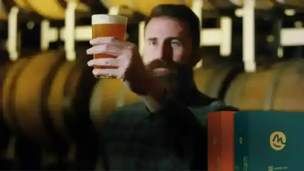 Uomo con barba esamina un bicchiere di birra dorata, con botti di legno sullo sfondo. Scatole di birre lager famose in primo piano.
