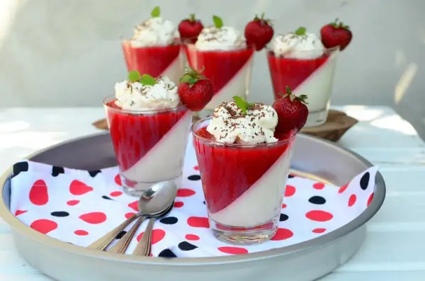 Panna cotta z truskawkami i bitą śmietaną w szklankach, udekorowana miętą i czekoladą.