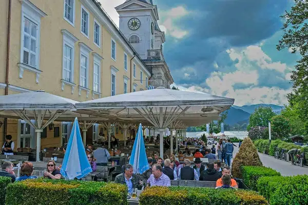 Menschen genießen die Tegernsee Feste unter Sonnenschirmen. Ein Gebäude mit Turmuhr und bayerische Fahnen im Hintergrund.