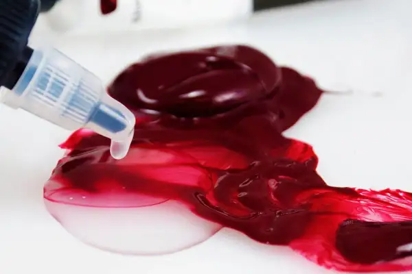 Un beccuccio trasparente rilascia un liquido chiaro su una macchia di vernice acrilica color bordeaux, creando sfumature vivaci.