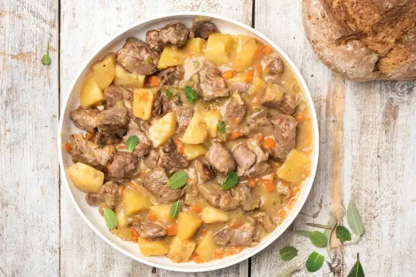 Un saporito agnello in umido con patate, carote e erbe aromatiche, servito in un piatto bianco su un tavolo di legno rustico.