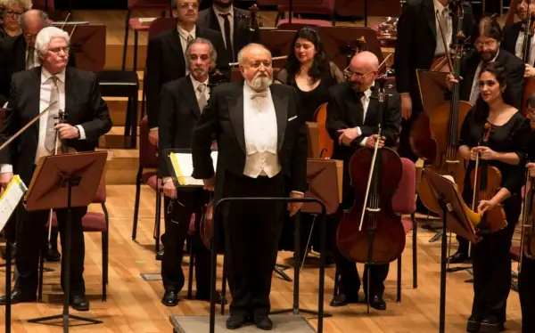 Orkiestra przygotowuje się do wykonania Dies Irae. Dyrygent w białej kamizelce i fraku stoi na podwyższeniu, otoczony muzykami z instrumentami smyczkowymi.