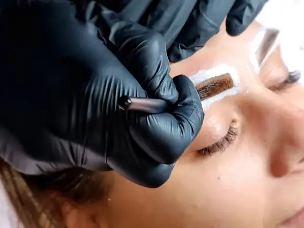 Stylist w czarnych rękawiczkach aplikuje hennę pudrową na brwi klientki.