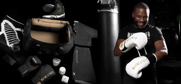 Floyd Mayweather Jr. smiles, holding a pair of white and gold Mayweather boxing gloves. The image also displays his boxing gear, including black gloves, shoes, and apparel.