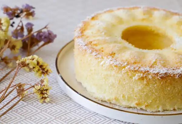 Delizioso chiffon cake ricetta originale americana, leggero e soffice, spolverato di zucchero a velo, con fiori secchi sullo sfondo.