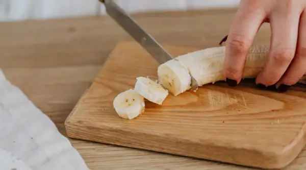 Eine Hand schneidet eine Banane in Scheiben auf einem Holzbrett. Perfekte Energie für das Training.