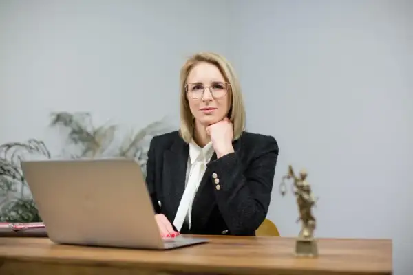 Kobieta w okularach i żakiecie siedzi przy biurku z laptopem i figurką Temidy.