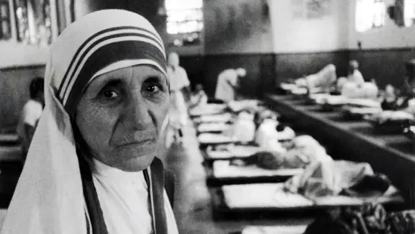 Mutter Teresa, deren Herkunft in Skopje liegt, blickt nachdenklich in die Kamera. Im Hintergrund liegen Kranke in Betten.