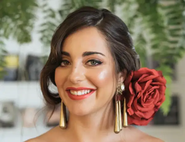 Peinados para feria pelo corto: mujer sonriente con rosa roja en el cabello, pendientes dorados y maquillaje festivo.