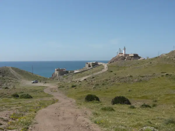Un camino de tierra lleva al restaurante El Faro Cabo de Gata, con el mar azul y un faro en la colina.