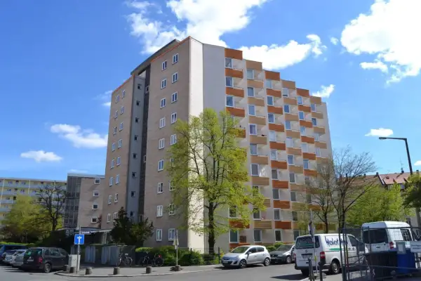Wysoki blok mieszkalny, który stanowi spadek po rodzicach, z balkonami i zielonym drzewem przed nim.