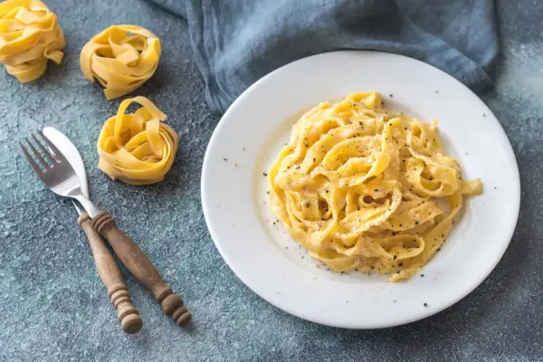 Un plat de fettuccine alfredo crémeux, saupoudré de poivre noir, avec des pâtes crues et une fourchette en bois à côté.