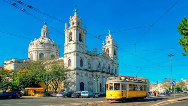 Un tranvía amarillo cruza frente a una iglesia blanca con cúpulas y torres, reflejando cómo se vive en Portugal con su encanto histórico y transporte icónico.