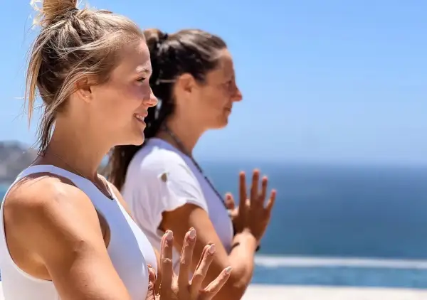 Dos mujeres practican yoga frente al mar, con las manos juntas en gesto de namasté.