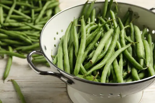 Haricots verts frais et humides dans une passoire, prêts pour la cuisson. Une pile de haricots verts est visible en arrière-plan.