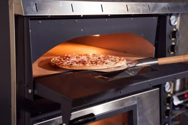 Une pizza garnie est en cours de cuisson dans un four professionnel, sur une pelle à pizza. La chaleur intense du four promet une cuisson parfaite.