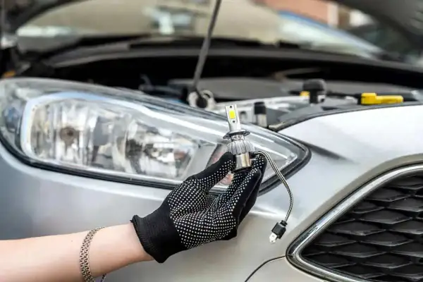 Ręka w rękawiczce trzyma nową żarówkę LED do wymiany przedniego reflektora w Peugeot 207.