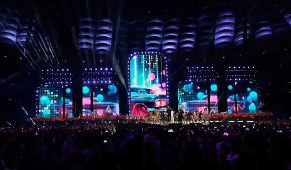 Wydarzenia na Stadionie Narodowym: scena z neonowymi samochodami i tańczącymi artystami, oświetlona przez reflektory. Tłum widzów wpatrzony w widowisko.