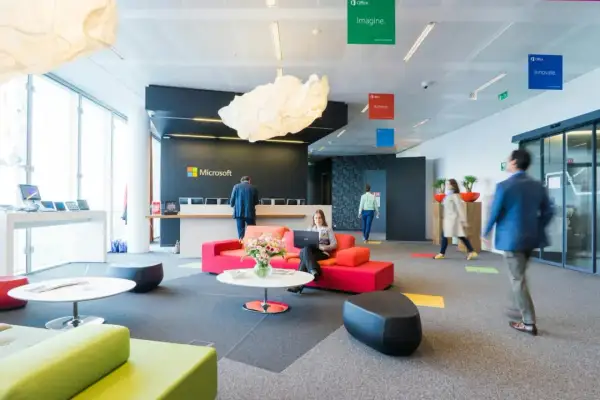 Un **open space** moderne chez Microsoft. Une femme travaille sur son ordinateur portable, entourée de canapés colorés. Des affiches "Imagine", "Achieve", "Innovate" flottent au plafond.