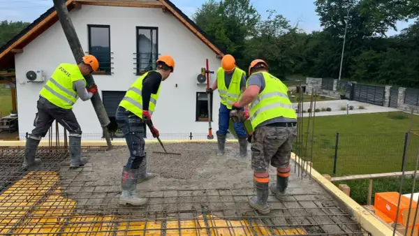 Budowlańcy w kamizelkach i kaskach, przestrzegając zasady bhp, wylewają beton. Jeden kieruje rurę, drugi grabi, trzeci wyrównuje.