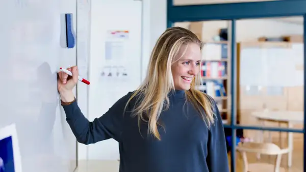Glad kvinna med blondt hår skriver på en whiteboard. Hon kanske studerar socialpedagog utbildning distans.