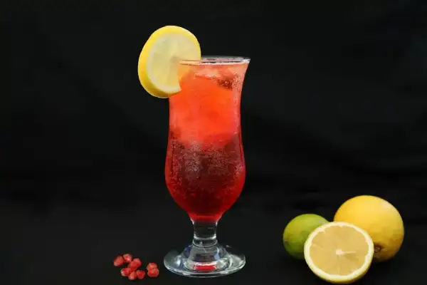Orzeźwiający drink z sokiem z granatu, ozdobiony plasterkiem cytryny. Obok leżą owoce granatu, cytryny i limonka.