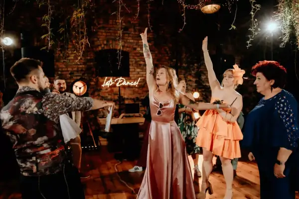 Oczepiny co to? Zabawa weselna w pełni! Panie w sukniach i panowie bawią się przy neonowym napisie "Let's Dance!".