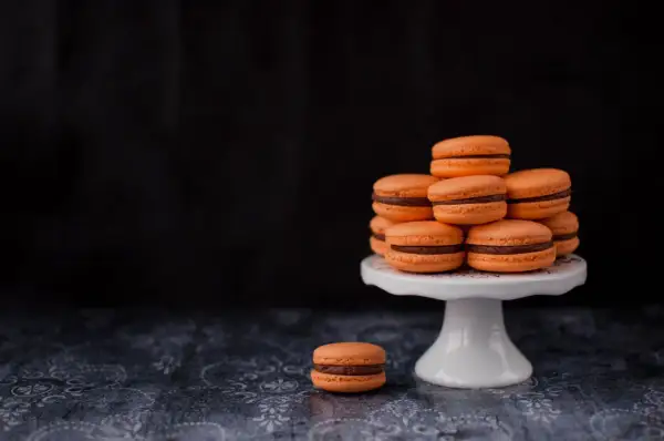 Pomarańczowe makaroniki francuskie z czekoladowym nadzieniem piętrzą się na białej paterze. Jeden makaronik leży obok.