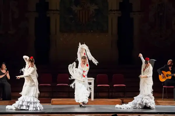 Mujeres en un apasionado baile flamenco, con trajes blancos y volantes, acompañadas por un guitarrista.