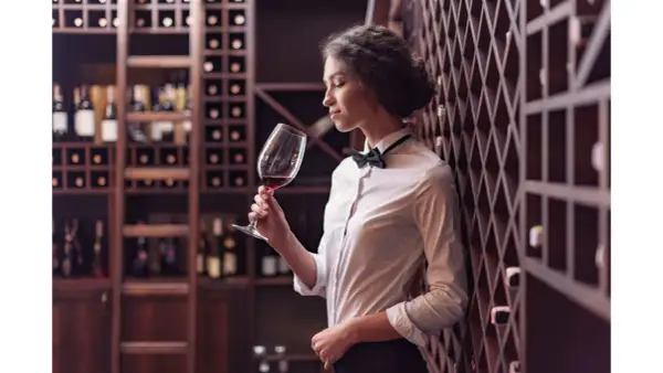 Elegancka sommelierka w białej koszuli i muszce wącha czerwone wino w kieliszku, otoczona regałami pełnymi butelek.