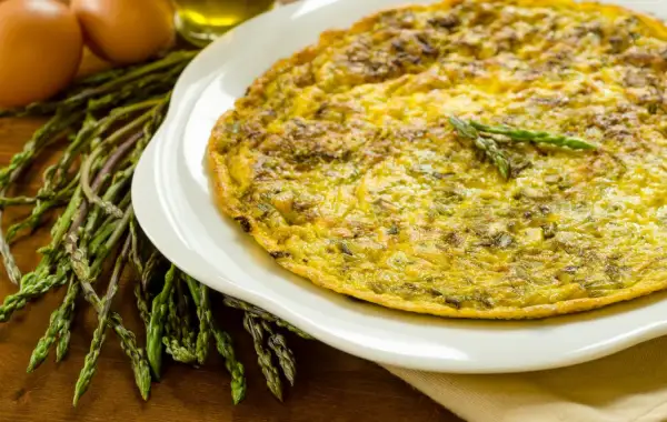 Frittata di asparagi selvatici dorata su piatto bianco, con asparagi freschi e uova sullo sfondo.