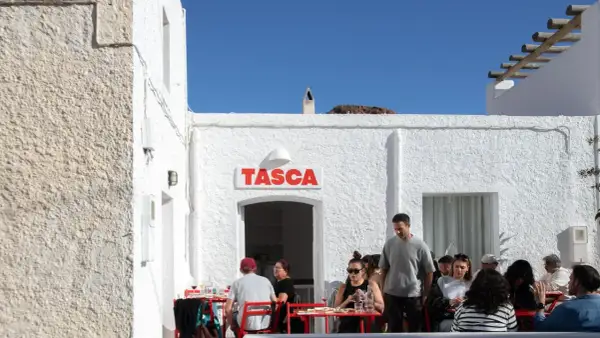 Gente disfrutando en el bar Tasca, con sus altas hortichuelas, bajo un cielo azul.