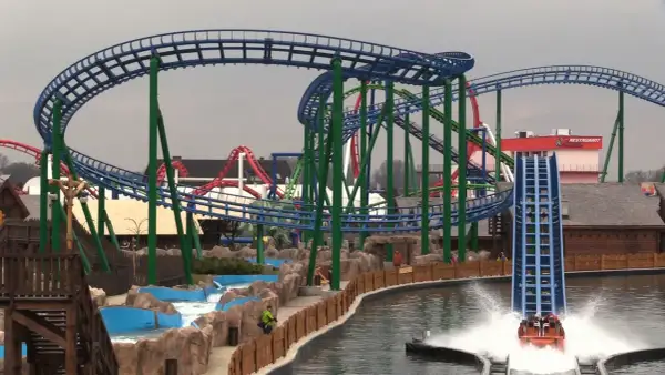 Water coaster zjeżdża z dużej wysokości, tworząc fontannę wody. W tle widać inne atrakcje parku rozrywki.