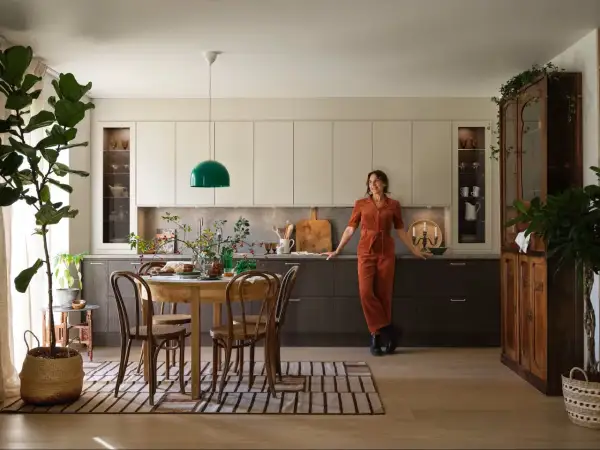 En kvinna i orange jumpsuit lutar sig mot köksbänken i ett kök med fransk inredningsstil. Ett runt träbord med stolar står framför köksöarna.