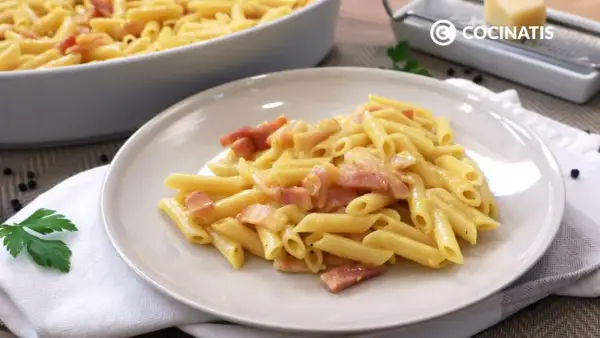 Plato de macarrones a la carbonara cremosos con trozos de panceta. En el fondo, un recipiente con más pasta y un rallador de queso.