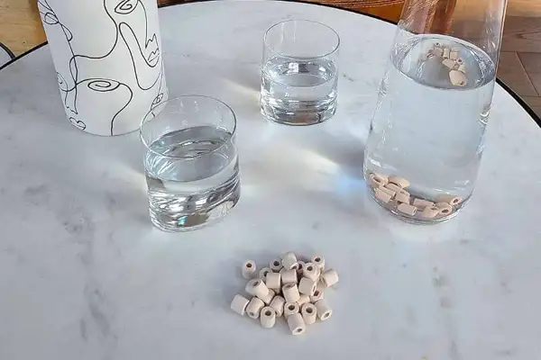 Des perles de céramique dans une carafe d'eau pour purifier l'eau du robinet naturellement. Des verres d'eau et un pichet décoratif complètent la scène.