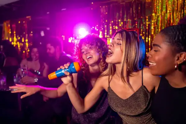 Trois amies chantent au micro lors d'une soirée karaoké à Paris. L'ambiance est festive, avec des lumières violettes et des guirlandes scintillantes.