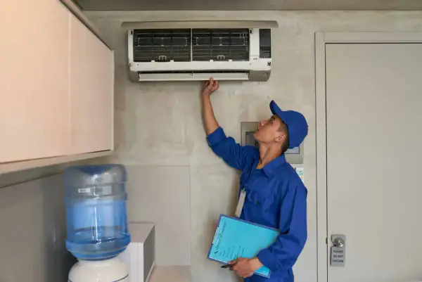 Un technicien en uniforme bleu vérifie une installation de climatisation murale, tenant un dossier de service.