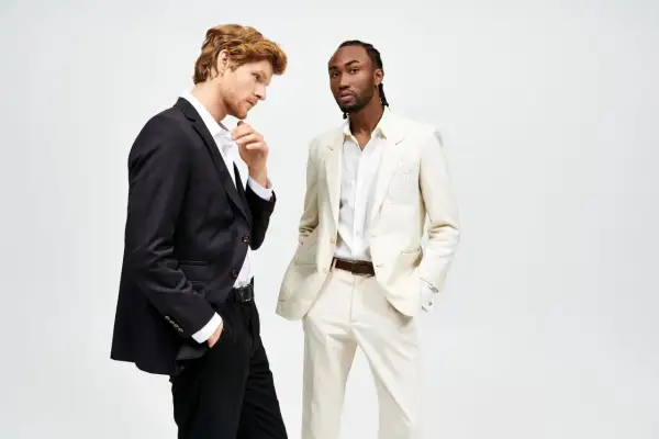 Due uomini in abiti eleganti. Uno con giacca nera e pantaloni neri, l'altro con giacca e pantaloni color crema. Il **slim fit significato** è evidente nella vestibilità moderna.
