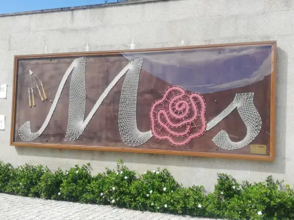 Letras de encaje blanco y rosa forman "Mos" en un letrero en el Pazo de Mos, con herramientas de encaje a la izquierda.