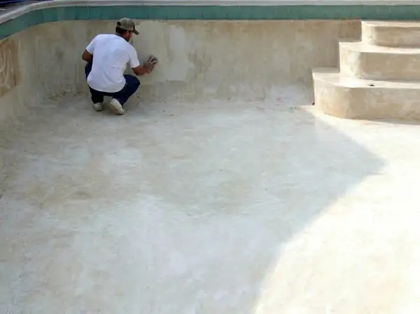 Hombre lijando la pared de una piscina vacía, preparándola para pintar cemento exterior.